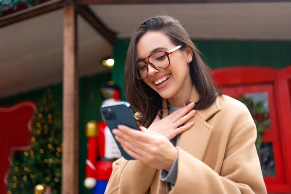 Surprised woman wear eyeglasses read positive message on smartphone. Happy female hold mobile phone in hand looking on device screen and smiles in surprise emotions. Christmas season online shopping
