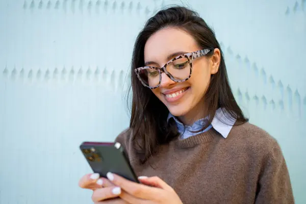 Official woman wear glasses smiles and looks at phone. Confident young adult woman use smartphone on bluew background. Modern female worker looks at workday schedule with smile