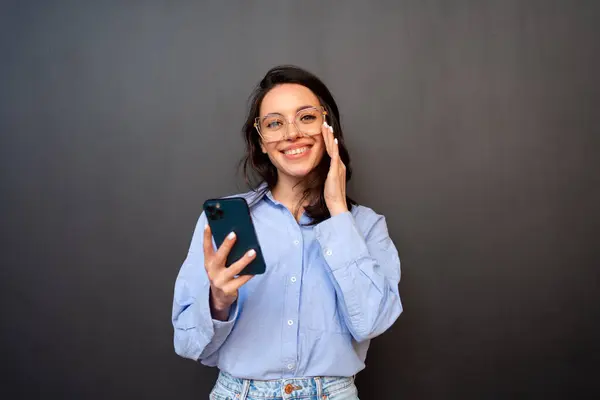 Smiling woman manager in formal wear standing isolated on black with mobile phone in hand. Girl in glasses, dressed shirt use smartphone