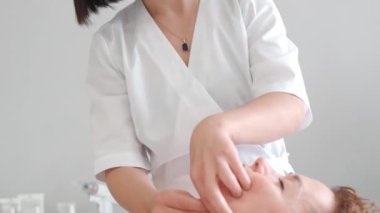 Kozmetik kabinesinde kadın, kafasına, omuzlarına ve boynuna masaj yapan masaj terapistinden yenilenen cilt bakımı uygulanıyor. Güzellik salonunda spa tedavisi ve masaj, dikey format.