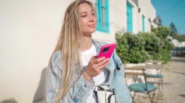 Avrupai bir kafede elinde akıllı telefonuyla oturan bir kadın, tipik Portekiz şehrinde yaz tatilinin tadını çıkarıyor. Restoran terasında cep telefonu kullanan mutlu, pozitif kadın turist.