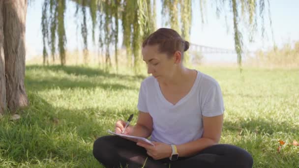 夏季的一天 一位女性形象地坐在公园的绿草上 双腿交叉 用笔在笔记本上写着便条 — 图库视频影像