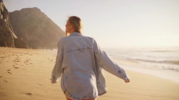 海のビーチを歩いて回り カメラに向かって微笑む女性 幸せな女性の旅行者はデニムシャツウォークサンディービーチ 夏の休暇を着た — ストック動画