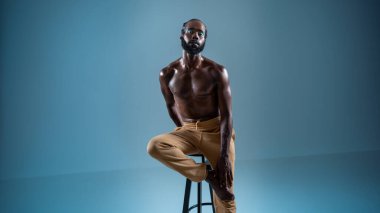 Siyah sakallı eşcinsel, mavi üzerine izole bir şekilde oturarak makyaj yapıyor. Afro-Amerikan eşcinsel, kaslı, tişörtsüz, makyajlı bir adam kameraya bakıyor.. 
