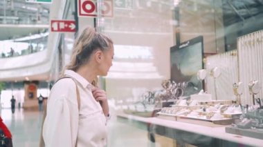 Orta yaşlı çekici bir kadın alışveriş merkezinde saatleri ve lüks mücevherleriyle dükkan vitrinine bakıyor. Kuyumcuda kendine hediye seçen beyaz sarışın kadın.