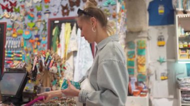 Genç bayan turistin hediyelik eşya dükkanında akıllı telefondan ödeme yaparken yan görüntüsü. Genç bir kadın Lizbon pazarını keşfediyor, kot gömlekli, tatil alışverişinin tadını çıkarıyor..
