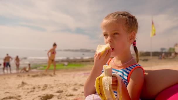 かわいいブロンドの女の子は砂浜で果物を食べています 夏休みに健康的な食事をする女性の子供 海岸近くで夏を満喫する飢えた少女 — ストック動画