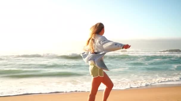笑顔の金髪の女性の肖像画は砂のビーチで手を伸ばしました 大きな岩と海の波の近くに立っているデニムジャケットとショートパンツの美しい女性 レディースは夏休みを楽しむ — ストック動画