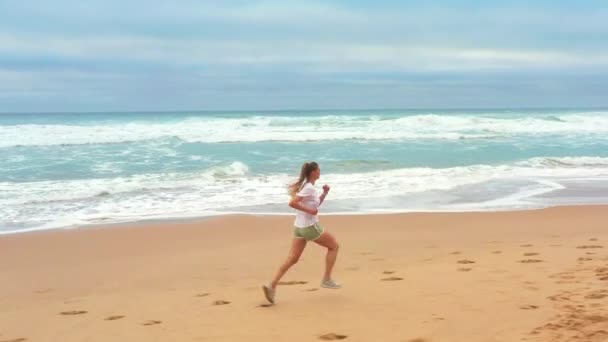 空中追跡ショット 夏の朝に美しいビーチでジョギングする若い女性 海岸を走る屋外で運動するアクティブな白人女性 健康的なライフスタイルとスポーツのコンセプト — ストック動画