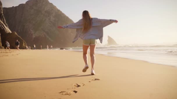ビーチを走る女性のバックビューデニムシャツ フライングヘア 日没のオーシャンビュー ビーチバケーション 旅行の概念の自由 海の夏休み 背景にある山々 — ストック動画