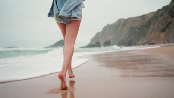 海で砂浜を走る若い女性のリアビュー デニムシャツの陽気な女性は 背景の山で濡れた砂の上を裸足で歩きます 幸せな女性は歩き回り そして休暇を楽しんでいます — ストック動画
