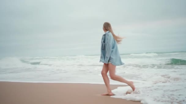 ブロンドの髪が走り 砂浜の海に沿って回転するケアフリーの女性の全長 夏休みのデニムシャツの女性観光客 エキサイティングな女性はビーチの休日にあります — ストック動画
