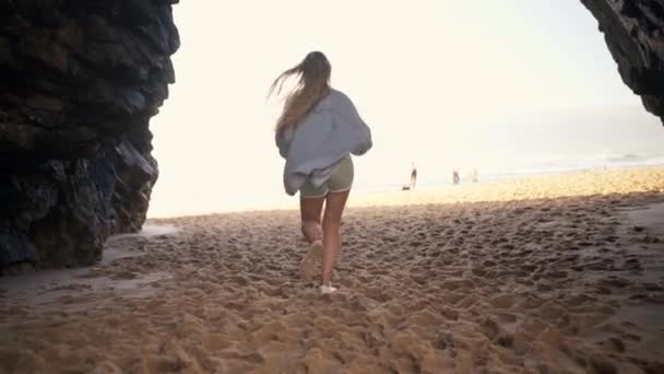 岩のなかで砂浜で走る陽気な若い女性のリアビュー デニムシャツと長いブロンドの髪を持つ遊び心のある女性は 晴れた日に砂浜で夏休みを楽しんでいます — ストック動画