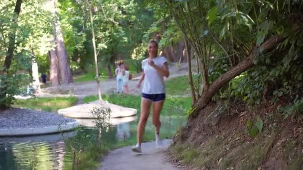 スポーティな若い女性ランナーは 夏の公園の池の近くの歩道でジョギングアクティブウェアで走ります 健康的なライフスタイルを表す自然の中で走るハッピージョギング — ストック動画