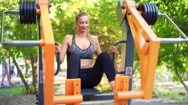 Spor elbiseli genç bir bayan gülümsüyor ve açık hava parkında tren simülatöründe bacak egzersizi yapıyor. Yaz parkında spor yapan kendine güvenen bir bayan sporcu. Sağlıklı yaşam tarzı kavramı.