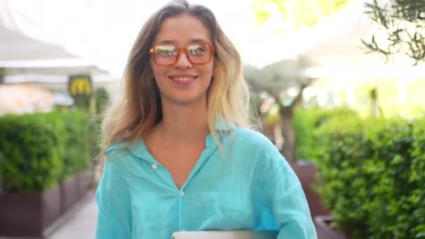 戴着时髦眼镜站在咖啡厅阳台上微笑的女人 漂亮的金发女人穿着蓝色衬衫 高兴地看着摄像机 绿地和街道花环背景的年轻女士 — 图库视频影像
