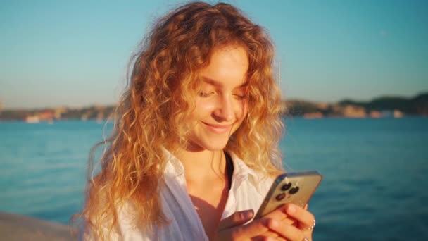 漂亮的年轻女子在河边用智能手机应用 在手机上用卷曲的金发发短信微笑的女人 社交媒体 连接概念 — 图库视频影像