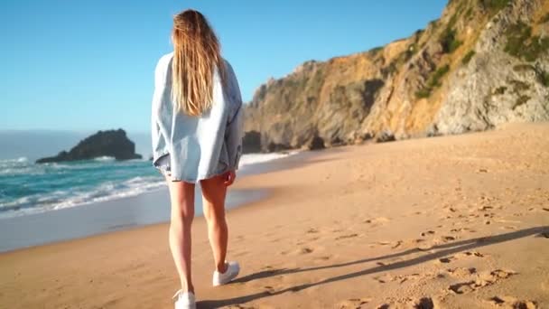海で砂浜を歩く若い女性のリアビュー デニムシャツの陽気な女性は バックグラウンドの山で夏休みを楽しんでいます ハッピーガールウォーク 晴れた日に風に回る — ストック動画