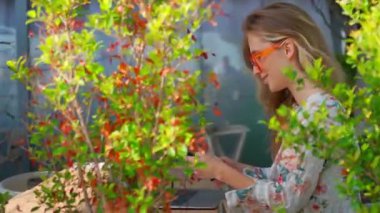 Akıllı telefonu dönen ve kameraya gülümseyen çekici genç bir kadının yan görüntüsü. Açık kafede oturan turuncu gözlüklü güzel bir kadın. Sarışın kadın şehrin bitkilerinin yanında oturuyor..