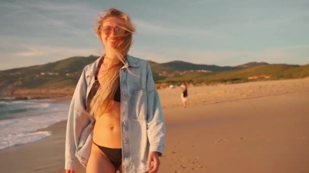快乐快乐的女人走在海边 背景是群山 穿着比基尼和斜纹棉布衬衫的活泼金发女游客旋转着 微笑着 看着镜头 在海滨度假 — 图库视频影像