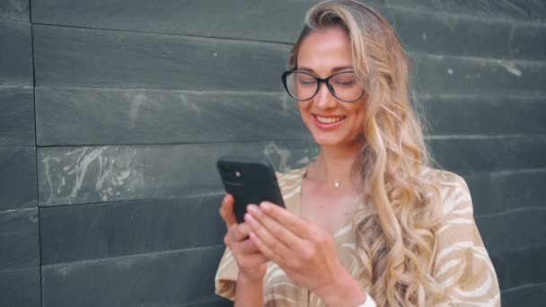 Videollamada Mujer Rubia Feliz Usando Teléfono Inteligente Apoyado Pared Gris — Vídeo de stock