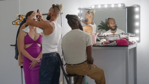 メイクアップアーティストは 照らされた鏡の前にひげ付きゲイモデルの近くの女性に髪型を作ります 男性と女性のパフォーマーは彼らの仕事のためのバックステージを準備します — ストック動画