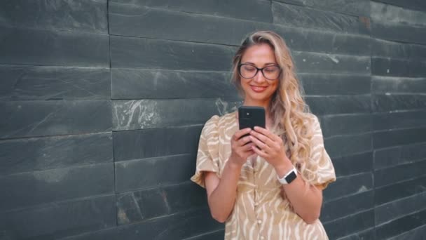 Mujer Sonriente Usando Teléfono Inteligente Apoyado Pared Gris Mujer Rubia — Vídeo de stock
