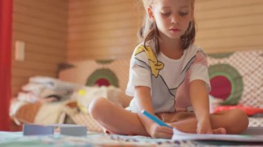 Düşünceli küçük beyaz kız elinde kalemle yatakta oturmuş defterine çizim yapıyor. Anaokulu çocuğu evde mektup öğreniyor. Bilgi ve evde çalışma. Çocukluk kavramı.