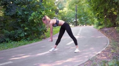 Spor kıyafetli bir kadın koşmadan önce ısınma egzersizi yapıyor. Parktaki asfalt yolda bacaklarını esneten aktif formda koşucu sağlıklı yaşam tarzını temsil ediyor. Gülümseyen uzun boylu sporcu kız..