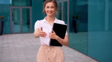 Ofis binasının dışında el sıkışma teklif eden belge klasörünü tutan kendine güvenen bir iş kadınının portresi. Anlaşmayı imzalamaya hazır gülümseyen genç bir kadın..