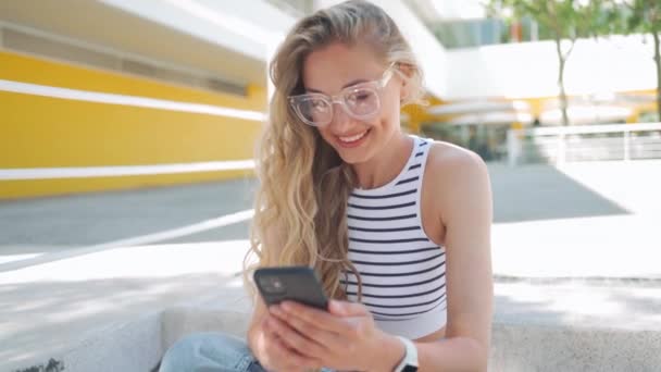 スマートフォンを使って眼鏡で笑顔の女性 外のステップに座っている携帯電話を介して幸せな美しい若い金髪の女性のテキストメッセージ オンラインショッピング ソーシャルメディア — ストック動画