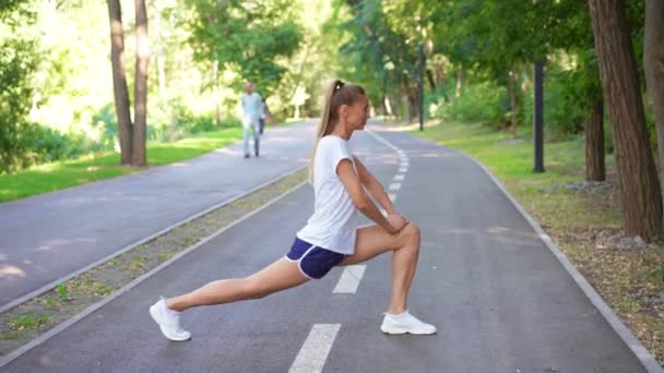 夏の公園で運動する前に足を伸ばす女性ランナー 若い運動女性がアスファルト道路で走る前に体を温めていると確信しています 女の子は健康的なライフスタイルを表しています — ストック動画