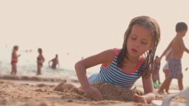 Yaz tatilinde plajda oynayan küçük kız, okyanus arka planında kalabalık turistlerle kumdan kale inşa ediyor. Yaz tatilinin tadını çıkar. Sahilde iyi eğlenceler.