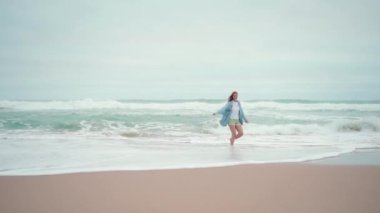 Okyanus sahilinde koşan ve kameraya gülümseyen mutlu bir kadın. Mutlu kadın gezgin kot gömlekli, kumlu deniz sahilinde, tatilde el kamerasıyla geziyor.