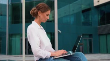 Modern ofis binasının dışında otururken dizüstü bilgisayar kullanan beyaz tişörtlü, gülümseyen genç iş kadınının yan görüntüsü. Şirket kadını netbook üzerinde çalışıyor. Şehirde esintinin tadını çıkarıyor..