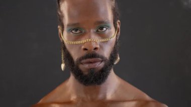 Siyah arka planda dans eden parlak makyajlı ve altın aksesuarlı kendine güvenen Afro-Amerikan sakallı eşcinsel. Stüdyoda yetişkin transseksüel gösterisi.