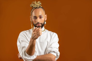 Sana ihtiyacımız var. Neşeli ve kendinden emin Afro-Amerikan gay, parlak makyajlı, kesin bir seçim yapıyor, kamerayı işaret ediyor, varyantlardan seçiyor, insanlarla konuşuyor, turuncu üzerinde izole edilmiş.