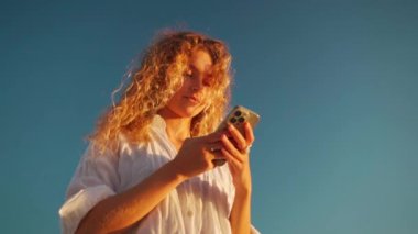 Akıllı telefon kullanan bir kadın sosyal medyayı kontrol ediyor. Kıvırcık saçlı sarışın kadın cep telefonu ekranına bakıyor. Mavi gökyüzü geçmişine karşı online uygulama kullanan genç bir kız. Düşük açı görünümü.