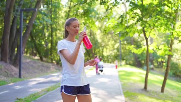 疲惫的年轻女子穿着运动服 慢跑后喝瓶子里的水和擦汗 在夏季公园的人行道上站着微笑的女运动员代表着健康的生活方式 — 图库视频影像