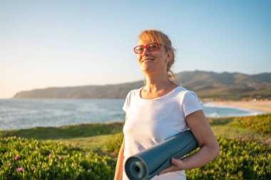 Yaşlı, beyaz, gözlüklü, yoga minderi tutan, kumsalda gülümseyen kadın. Okyanus kıyısında sabah yoga seansına hazırlanmak, sağlıklı yaşam tarzını desteklemek ve tatilde emekliliğin tadını çıkarmak.