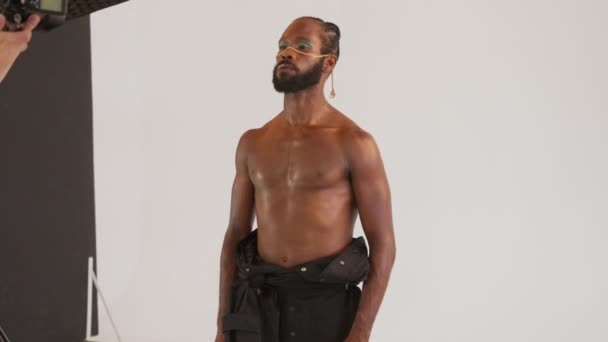 マッサージシャツレスのアフリカ系アメリカ人のゲイ男性は 白い背景に対してスタジオでプロのカメラマンのためにポーズを構成します Lgbtqを撮影する写真家 — ストック動画