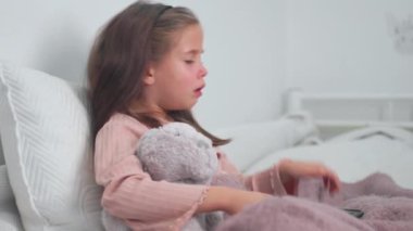 Evde yatakta yumuşak bir oyuncakla otururken öksüren hasta bir kız. Soğuk algınlığı ve grip geçiren üzgün bir kız ve dairede dijital tablet kullanıyor..