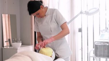 Üniformalı kadın kozmetik uzmanı müşterisinin yüzüne sarı aljinat maske takıyor güzellik salonunda rahat bir yatakta yatıyor..