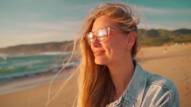Kumsalda rüzgarlı saçları olan gülümseyen bir kadın. Gözlüklü ve kot gömlekli güzel bir bayan turist gözlerini kaçırıyor. Plaj tatili.