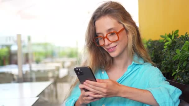 アウトドアカフェでスマートフォンを使用したブルーシャツの笑顔の女性 携帯電話の長いブロンドの髪の読書テキストメッセージを持つ眼鏡の若い女性 モバイルデジタルデバイスを使用したオンライン通信 — ストック動画