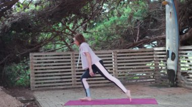 Kadın, çam ormanında, tahta zeminde yoga yapıyor. Orta yaşlı kadın savaşçı pozu yavaş çekimde meditasyon yapar. Zindelik kavramı, sağlıklı yaşam tarzı