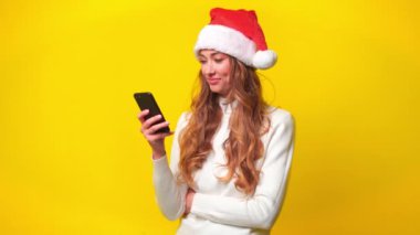 Akıllı telefon kullanan ve sarı arka planda el kol hareketi yapan heyecanlı bir Noel kadını. Neşeli genç bayan arka planda cep telefonu kullanırken baş parmağını kaldırıyor.. 