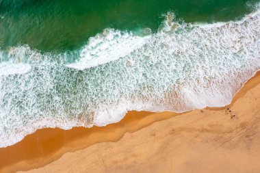 Kumlu sahil şeridinde dalgalanan turistik deniz dalgalarının güzel, pürüzsüz görüntülerini gösteren drone görüntüsü. Altın plajın havadaki görüntüsü derin mavi okyanus suyu ve köpüklü dalgalarla buluşuyor.
