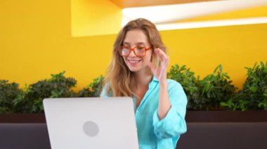 Neşeli kadın, kafeterya masasında dizüstü bilgisayar kullanarak gözlük ayarlıyor. Mavi gömlekli, uzun sarı saçlı, kahve dükkanında çalışan mutlu bir kadın. Genç bayan kameraya gülümsüyor..