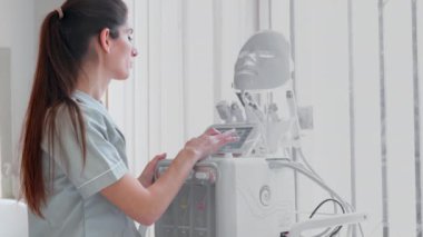 Modern klinikte modern hidroterapi makinesi kullanan kadın kozmetikçi. Genç beyaz profesyonel, kuaförde tedaviye hazırlanırken ekipman kuruyor ve iyontoforez kablosu seçiyor.. 